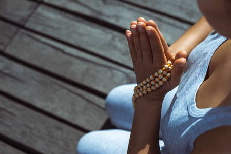 The young woman sitting on a wooden pier with her hands folded with a rosary in a prayer pose. Meditation, yoga in nature. Close-up. View of the hands.の写真素材