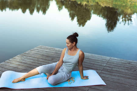 The young woman sits on a mat in a free pose by the lake. Yoga class in nature.の写真素材