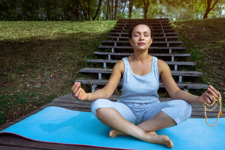 The young woman sits on a blue mat on a wooden pier in a lotus position with a rosary in her hands....