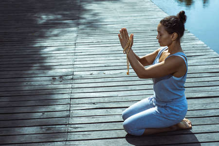 The young woman is sitting on a wooden pier with her hands folded in a prayer position on her head. General side view. Meditation, yoga in nature.の写真素材