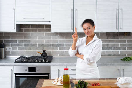 The young woman chef stands in the kitchen in front of the table holding her thumbs up.の写真素材