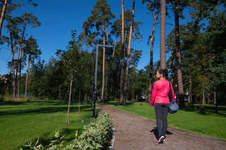 The young woman with a sports bag on her shoulder walks along the path in the park. Back view.の写真素材