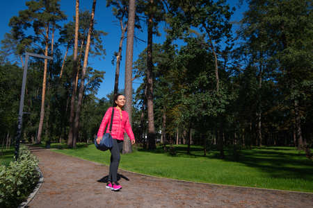 The young woman with a sports bag on her shoulder is walking along the path in the park. Front view.の写真素材