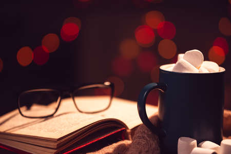Still life. The cup with marshmallows on a background of colored lights. In the background, an open book and glasses.の写真素材