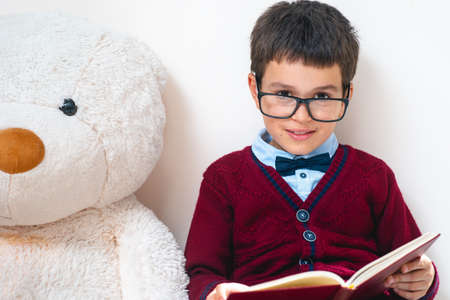 The schoolboy in a sweater and glasses sits next to a large teddy bear with an open book in his hands and looks at us. Conceptual. Copy space.の写真素材