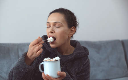The young woman eats a spoonful of marshmallows from a white mug. Drinking hot chocolate.の写真素材