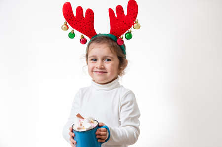 The girl with a hoop in the form of deer antlers stands on a white background with a cup of marshmallow in her hands. Christmas concept.の写真素材