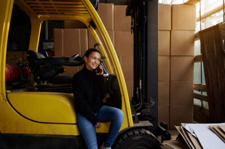 Inside the warehouse, the young woman businessman sits on the step of a yellow forklift and speaks on the phone. The sun is shining through the warehouse windows.の写真素材