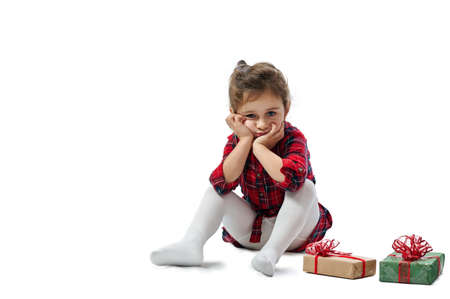 Isolated portrait of the child on white. A girl in a red dress sits with her face in her hands, next to Christmas gifts.の写真素材