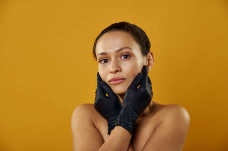 The beautiful african woman in black latex gloves touching face isolated on yellowの写真素材