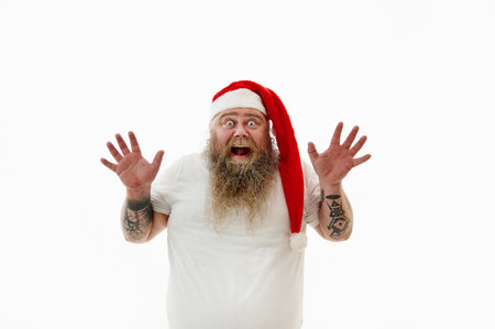 Isolated portrait on a white background of an overweigh man in Santa Claus hat raising palms and has surprised looks. Man with tattoed arms raises palm expressing surprised emotionの写真素材