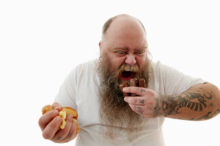 An overweigh caucasian bearded man in white t-shirt consuming unhealthy but delicious junk food. Wildly looking at chocolate barsの写真素材