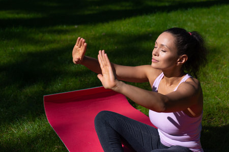 Young woman sitting on a fitness mat on a green grass in the park enjoying sunny day while...