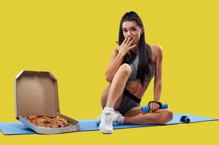 Fit woman covering mouth with her hand while sitting on a fitness mat near a box with tasty pizza and holding a dumbbell in the other hand. Isolated portrait on a yellow backgroundの写真素材