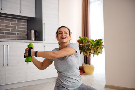 Smiling woman stretching her body with dumbbells after workout training at home. Workout during...