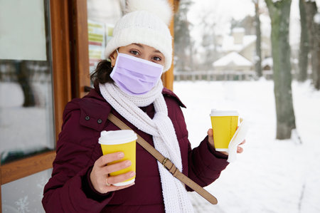 Brunette woman wearing medical mask holding two takeaway yellow cups of coffee or tea
