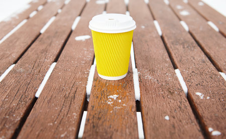 Yellow takeaway cup of hot drink, coffee or tea on wooden bench in a winter parkの写真素材