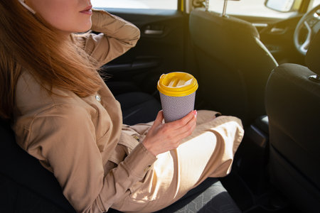 Young woman sitting on the rear passenger seat of a car holds a cup of tea or coffee and listens to the music. Cropped imageの写真素材