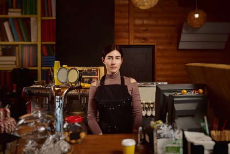 Portrait of a brunette woman barista in apron standing behind the bar