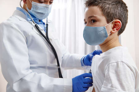 Female pediatrician in medical uniform uses a stethoscope while auscultating a little boy in protective medical maskの写真素材