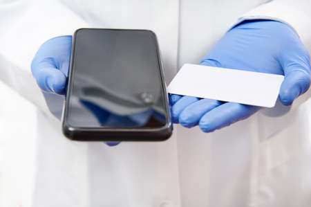 Cell phone and plastic white card on hands in medical gloves on the background of doctor's white uniform.の写真素材