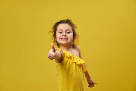 Beautiful little girl shows thumb up to the camera and smiles while posing on a yellow backgroundの写真素材