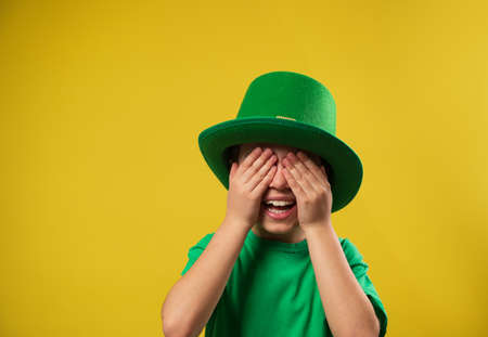 Laughing boy in green Irish leprechaun hat covers his eyes with his palms. Saint Patrick Day on yellow background with copy spaceの写真素材