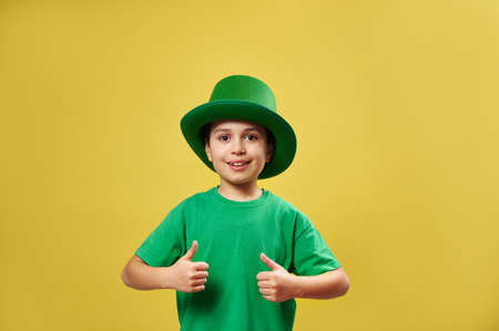 Smiling little boy in Irish green leprechaun hat showing thumbs up at camera standing on yellow background with copy space. Saint Patrick Dayの写真素材