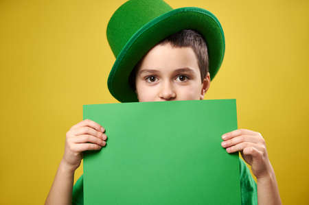 Handsome boy dressed in a leprechaun with a green Irish cap covers his face with a sheet of green paper. Saint Patrick's Day concept.の写真素材