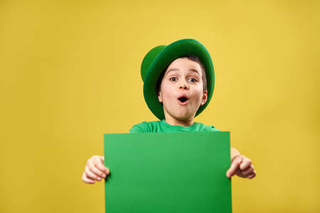 Surprised little boy in green Irish leprechaun hat poses wit a green sheet of paper in his hands. Saint Patrick's Day concept. Yellow background. Copy space.の写真素材