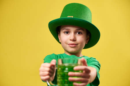 A boy wearing a green leprechaun hat holds a glass of green drink and holds it out in front of him to the camera. Celebrating Saint Patrick's dayの写真素材