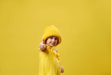 Beautiful little girl shows thumb up to the camera and smiles while posing on a yellow background with copy spaceの写真素材