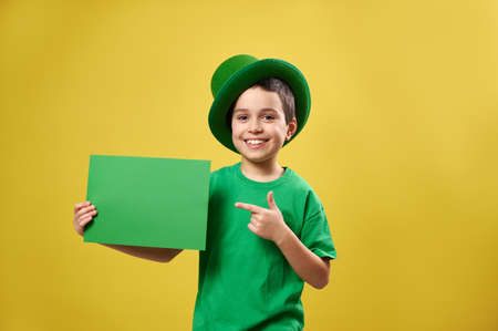 Happy boy in green clothes and Irish hat puts his index finger to a sheet of paper with copy space and smiles while posing on a yellow background. St Patrick's Dayの写真素材