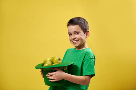 Smiling boy holding a leprechaun hat full of green apples. Saint Patrick's Day concept. Copy space. Yellow background.の写真素材