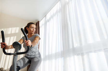 Young fit woman using stationary bike for cardio workout at home. Healthy and sporty lifestyle...