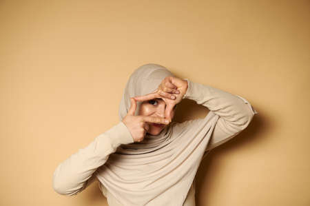 Beautiful muslim women in hijab making a frame with fingers and looking through it. Portrait on beige background with copy space. Facial emotions.の写真素材