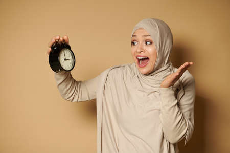 A stunned muslim woman wearing hijab holds an alarm clock in her hand and looks at it in horror because of fleeting time. Beige background with copy space.の写真素材