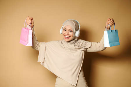Muslim Arab woman with covered head in hijab raises her arms up with bags in them and cute smiles to the camera with beautiful toothy smile. The human emotions, facial expression concept.の写真素材