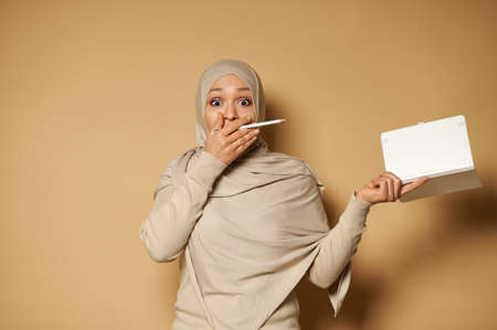 Surprised woman in hijab holds a note pad and pencil in her hands and cover her mouth expressing confusion, surprise and astonishment. Beige background with copy space.の写真素材