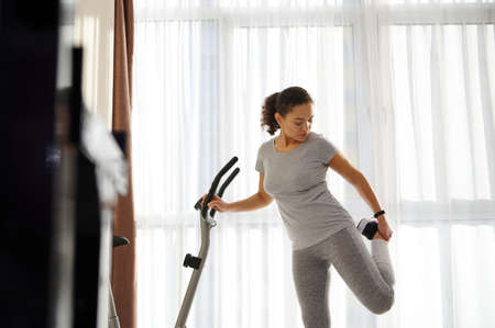 Young woman in gray sportswear stretching the muscles of the legs after training at home learning...