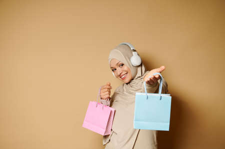 Adorable arabic muslim woman in hijab with a beautiful toothy smile stands sideways to the camera and posing holding out a gift bag on a beige background with copy spaceの写真素材