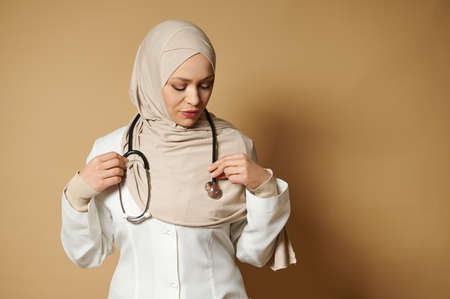 Confident portrait of a muslim woman doctor wearing hijab and putting a phonendoscope around her neck. Beige background with copy spaceの写真素材