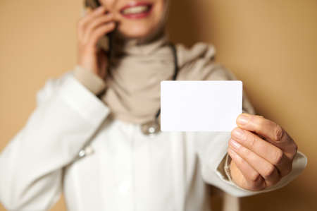 Selective focus on a plastic blank white card on the hand of a female Muslim doctor wearing a hijab talking on the phone. Copy space for promotions. Advertising. Insurance conceptの写真素材