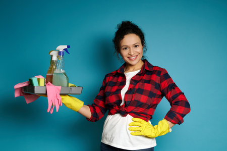 A charming brunette with curly hair, dressed in a white T-shirt, a red check shirt and yellow rubber gloves, holds a tray of cleaning supplies smiles while posing on a blue background with copy spaceの写真素材