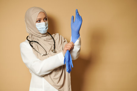 Muslim female doctor with covered head wearing medical protective gloves posing in front of camera on beige background with copy spaceの写真素材