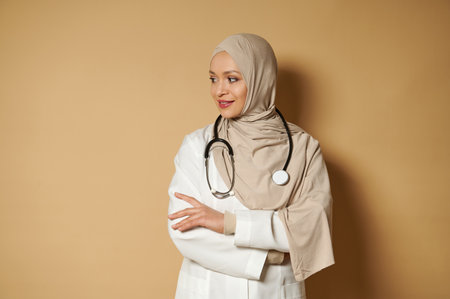 Attractive serene Muslim doctor with stethoscope around her neck in hijab standing with crossed arms and looking side on beige background with copy spaceの写真素材