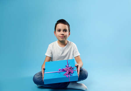 Cute smiling boy posing with a birthday gift in his hands on blue background with copy space. Happy birthday concept.の写真素材