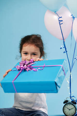 Focus on blue gift with a pink bow, which a cute little girl holds out to the camera while sitting on a blue background. Celebration concept. Happy childhood.の写真素材