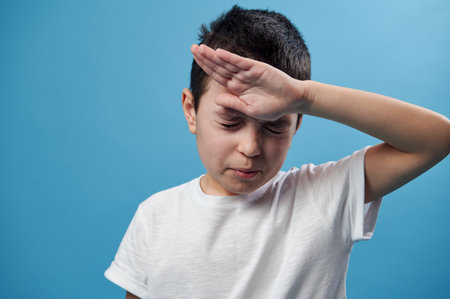 Boy holding his temple. Facial emotion. Closeup on blue background with copy space.