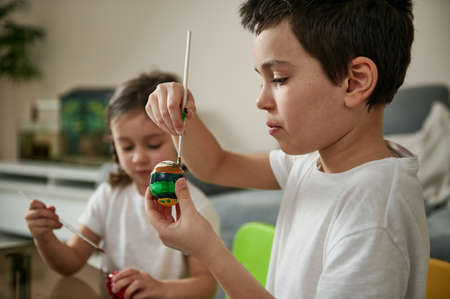 The schoolboy focused on decorating the Easter egg with a brush and colored paints while sitting at...